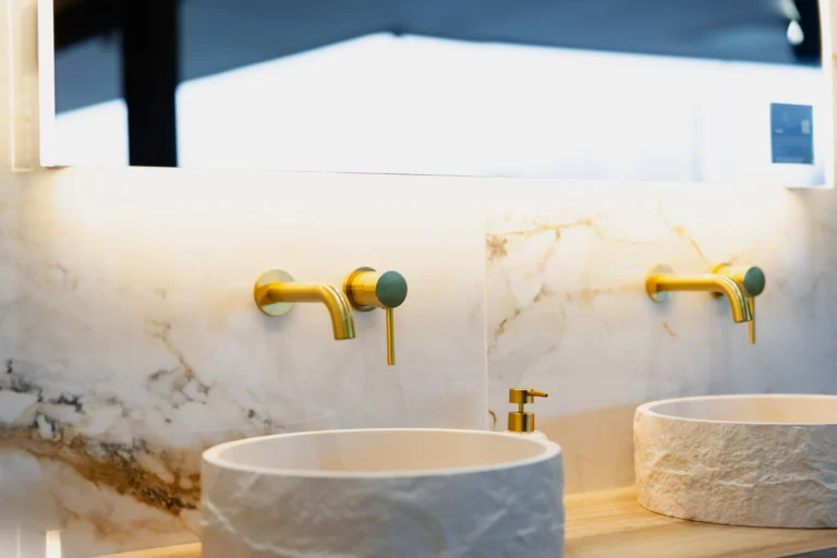 Brushed brass wall-mounted basin taps with sage-green handles above two textured vessel basins, against gold-veined marble-effect tiles