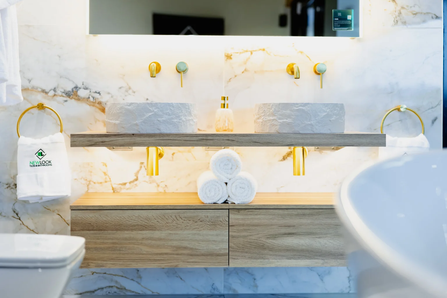Two textured white vessel basins on an oak shelf with brushed brass wall-mounted taps, set against white marble-effect tiles veined in gold and grey