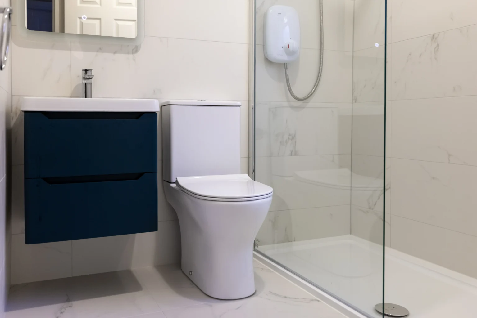 Bathroom with white marble-effect wall and floor tiles, a navy wall-hung vanity unit, a white toilet and a low shower tray behind a glass screen