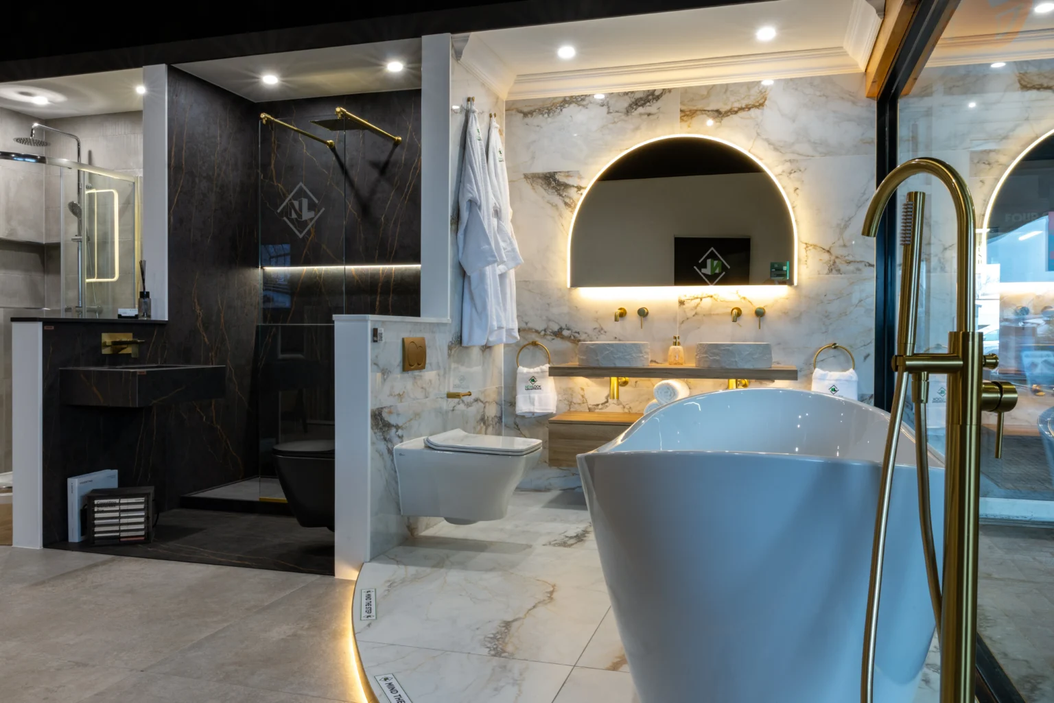 Showroom bathroom with white marble-effect walls and floor veined in gold, a freestanding bath with a brushed brass floor-standing tap and an arched backlit mirror