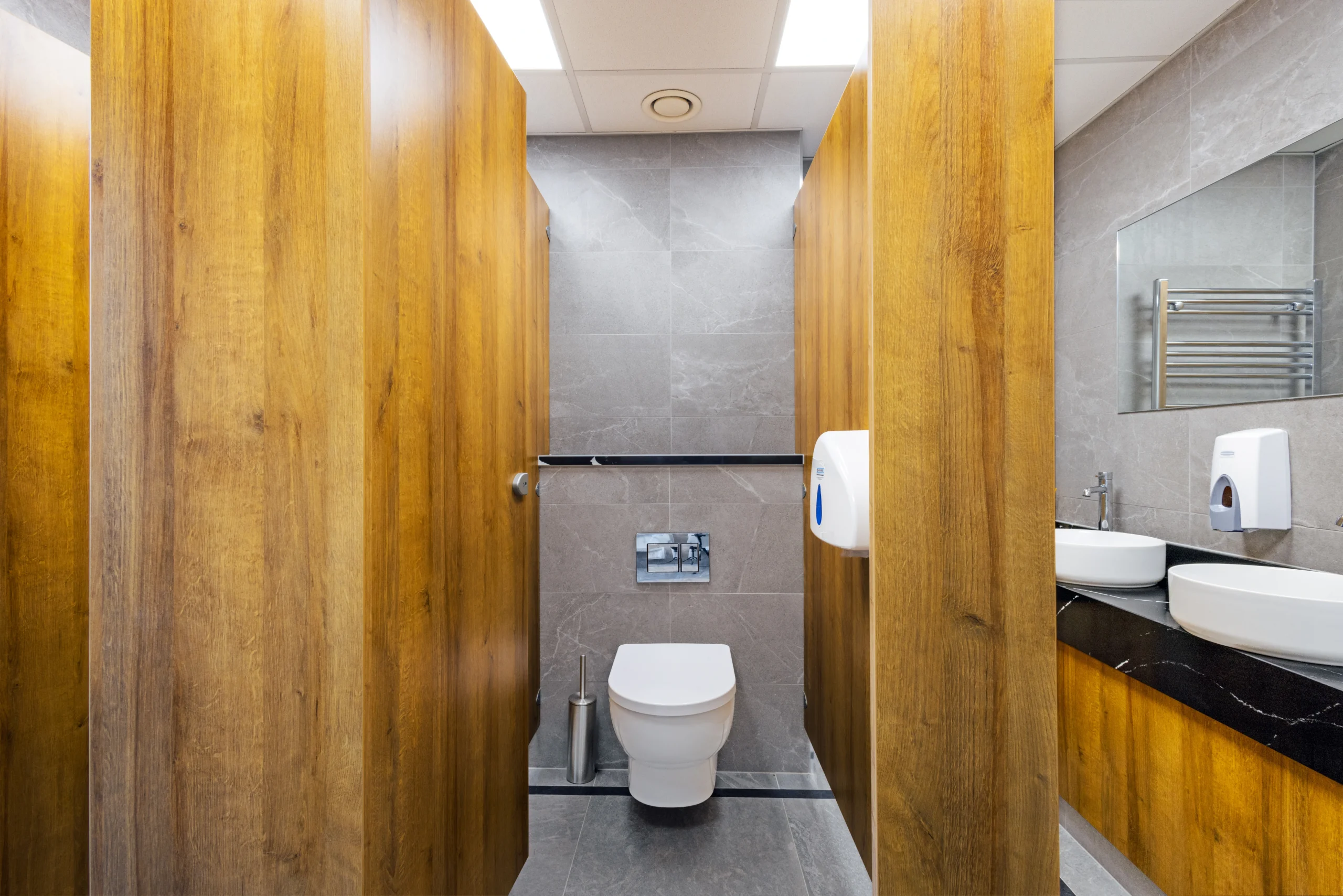 Sutton Golf Club commercial gents cubicle view — rimless wall-hung WC, chrome flush plate, large-format grey stone-effect porcelain tiles on walls and floor, flanked by floor-to-ceiling oak-effect cubicle panels.
