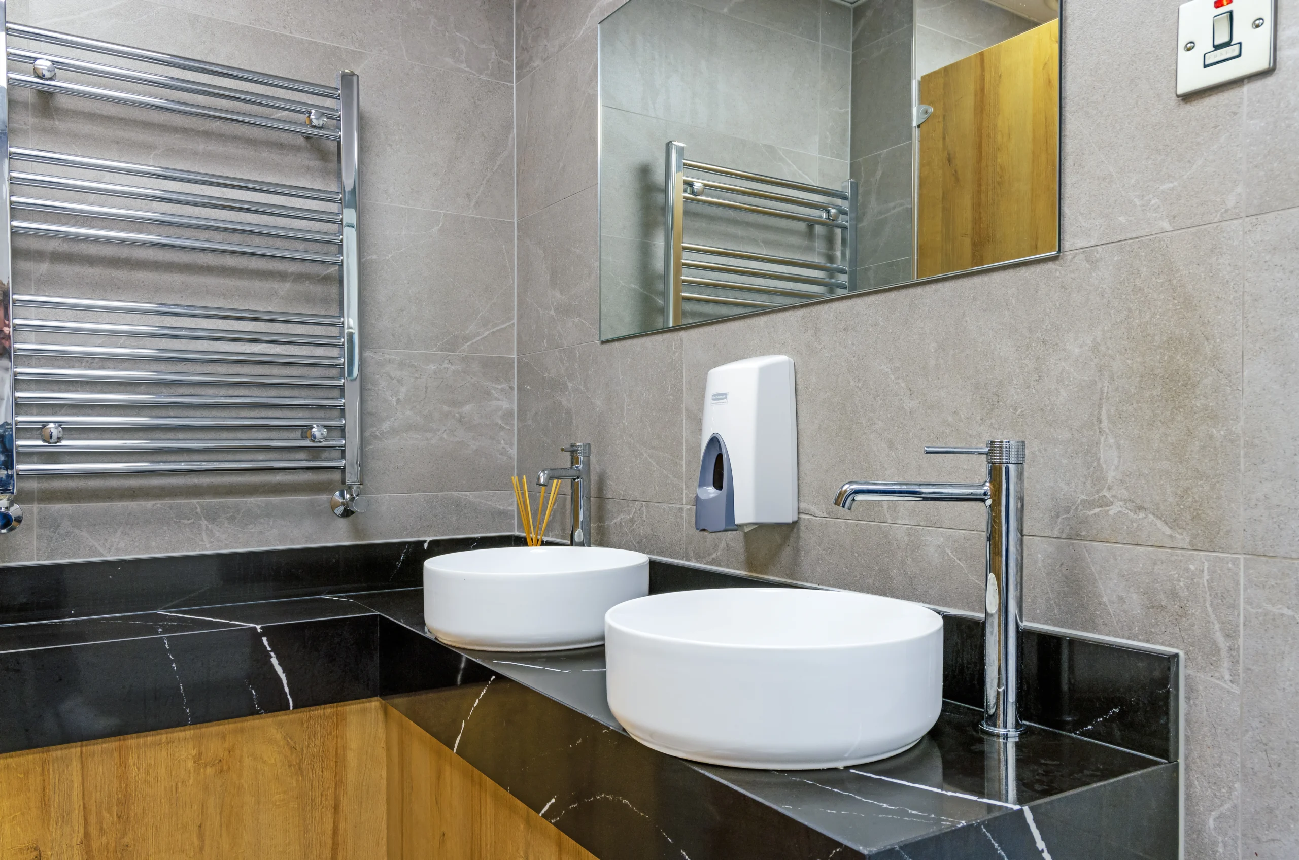 Twin-basin vanity in the gents at Sutton Golf Club — black veined marble-effect worktop, two round white counter-top basins, tall chrome single-lever mixer taps, mirror above and a chrome ladder-style heated towel rail on grey porcelain wall tiles.