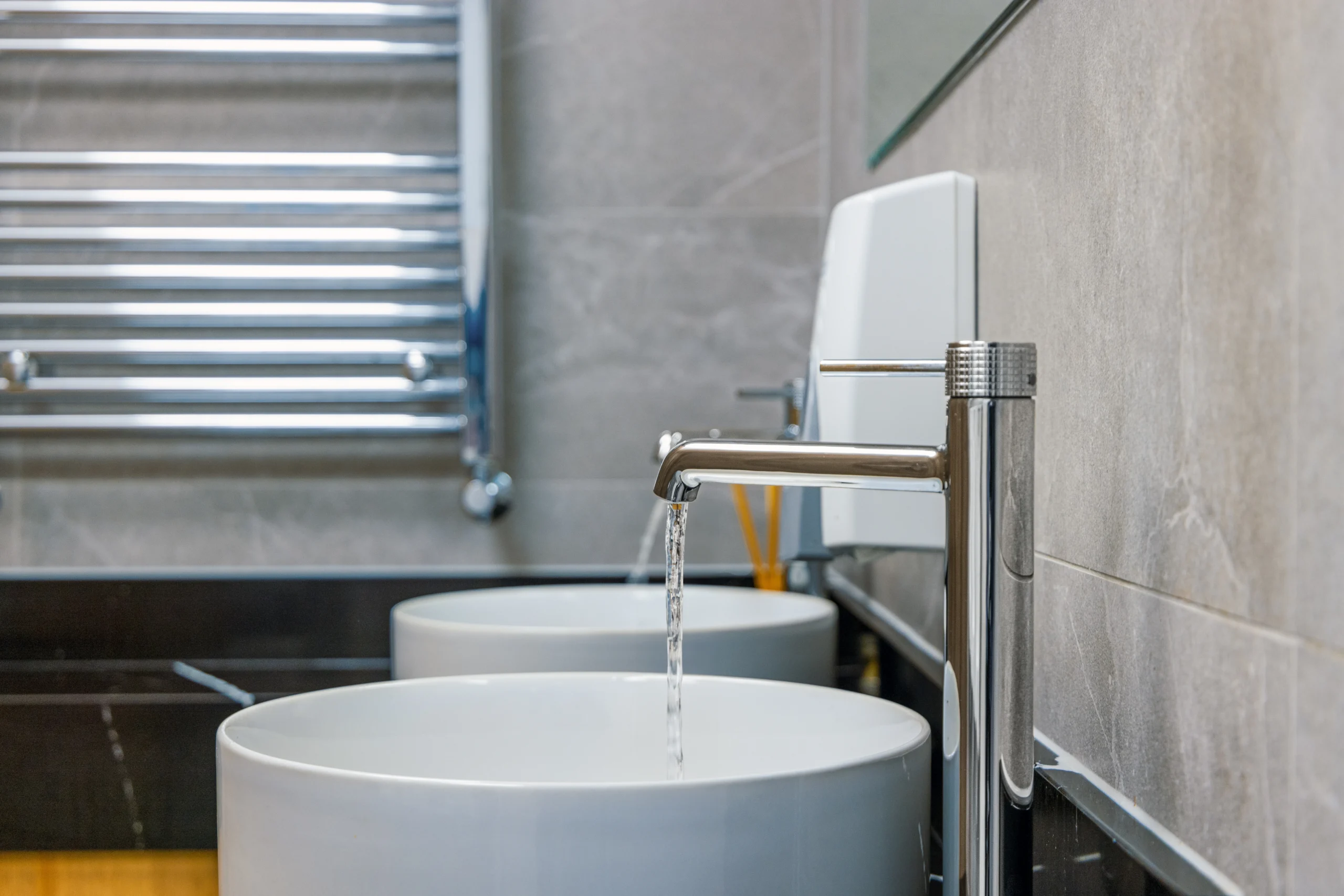 Detail shot of a tall chrome vessel-basin mixer in the gents washroom at Sutton Golf Club, water flowing into a round white counter-top basin on a black veined marble-effect worktop.