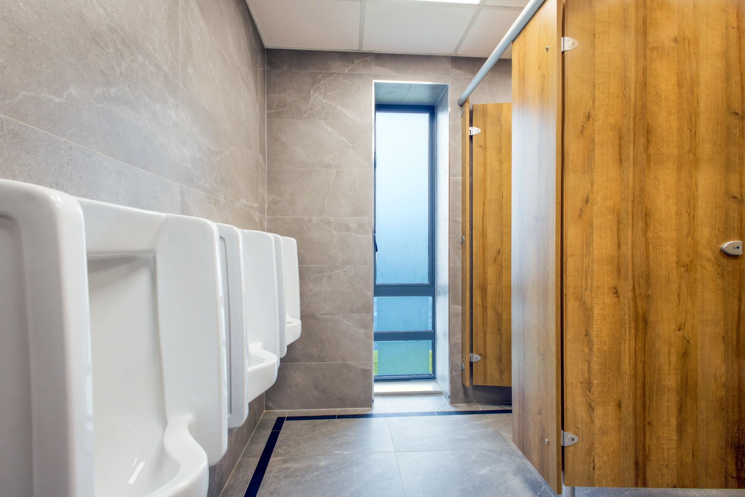 Urinal bay at Sutton Golf Club first-floor gents commercial bathroom — a row of wall-hung white ceramic urinals on large-format grey stone-effect porcelain, with oak-effect cubicle doors to the right.