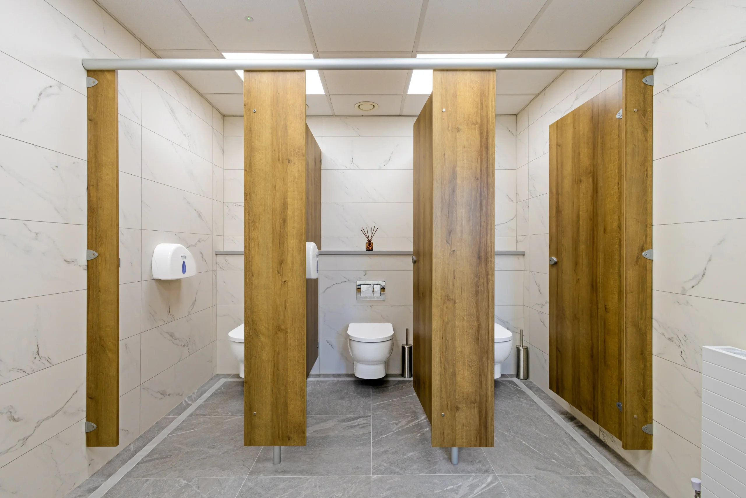 Sutton Golf Club first-floor ladies washroom — three oak-effect WC cubicles with white veined marble-effect porcelain walls from floor to ceiling, grey stone-effect porcelain floor and wall-hung toilets.