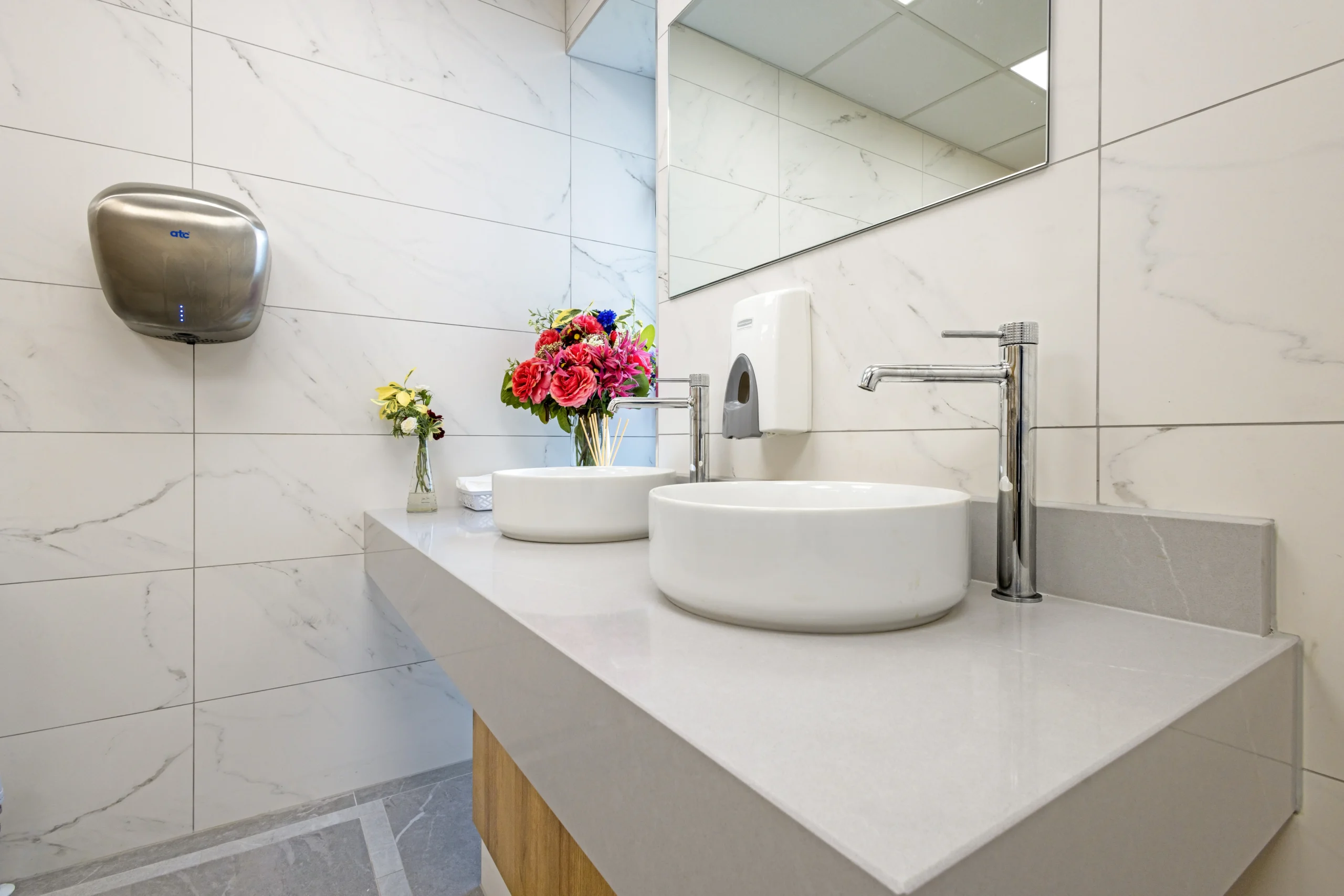 Detail of the ladies vanity at Sutton Golf Club — round white counter-top basin, tall chrome single-lever mixer, white veined marble-effect porcelain wall backdrop and a stainless steel hand dryer to the side.