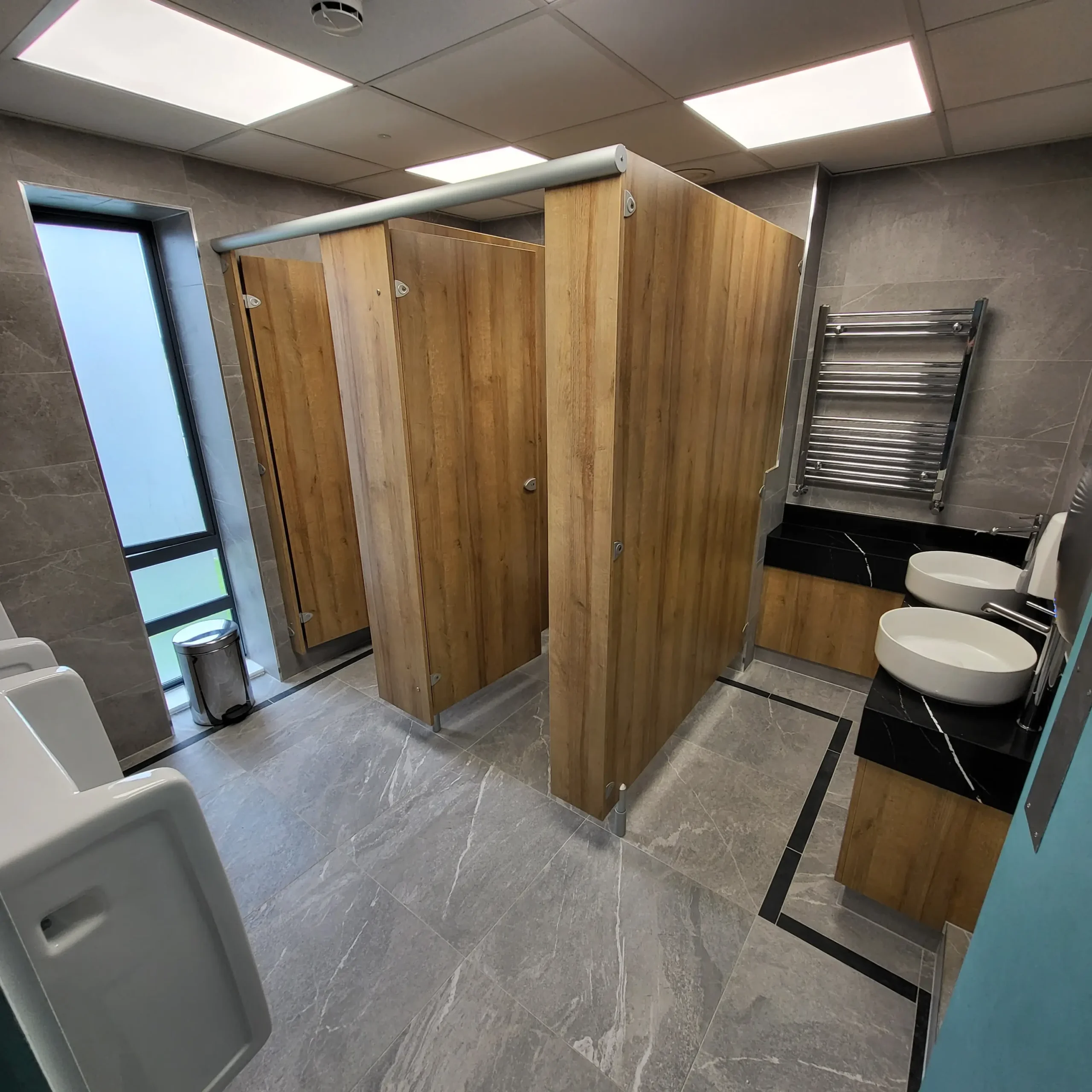 First-floor gents commercial washroom at Sutton Golf Club — wide view showing the urinal bay to the left, oak-effect cubicle block in the centre, and twin-basin vanity with chrome heated towel rail to the right, all on large-format grey stone-effect porcelain.
