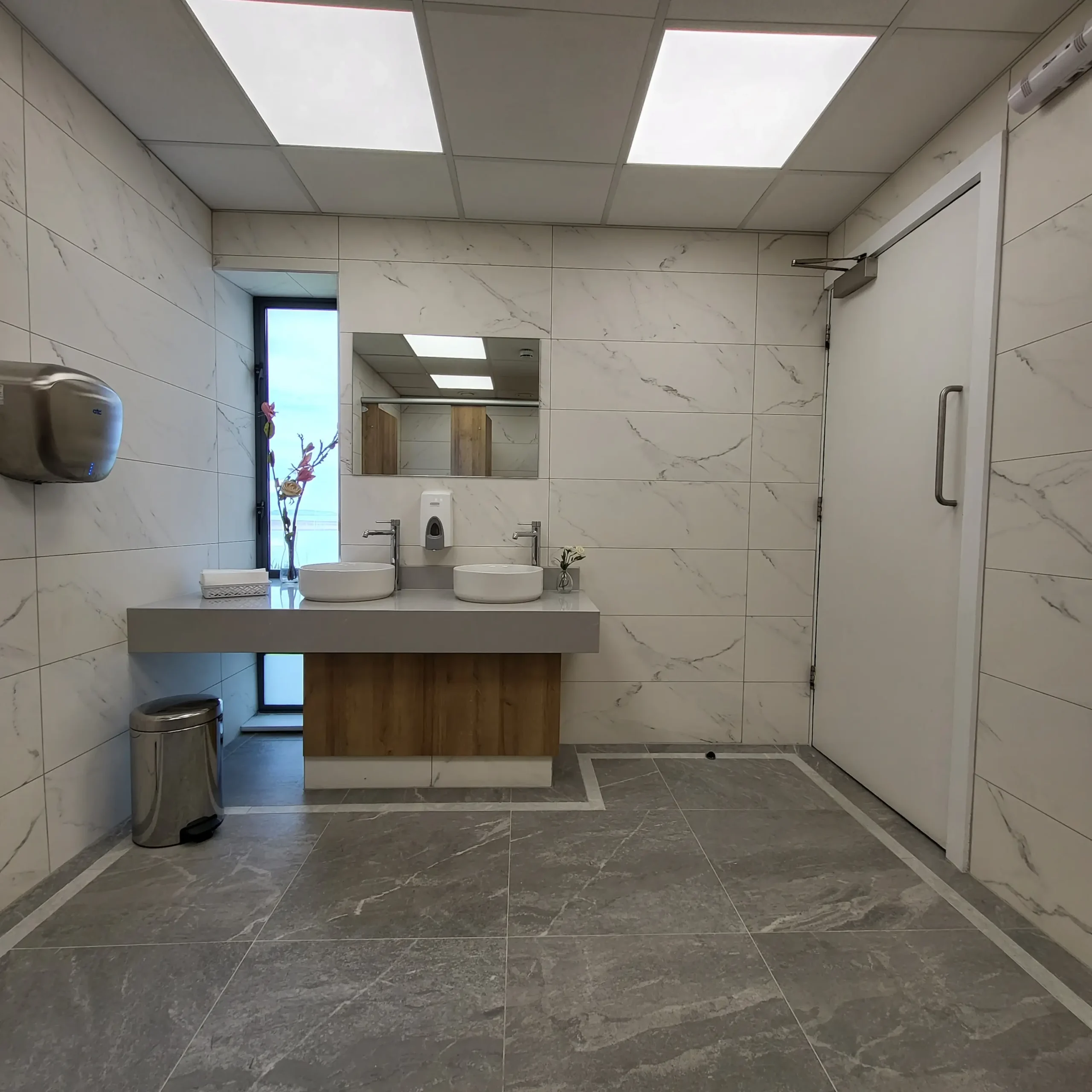 First-floor ladies washroom at Sutton Golf Club — wide view with a bespoke solid-surface vanity on an oak base unit, frameless mirror and twin round basins, framed by white veined marble-effect porcelain walls and a grey stone-effect floor.