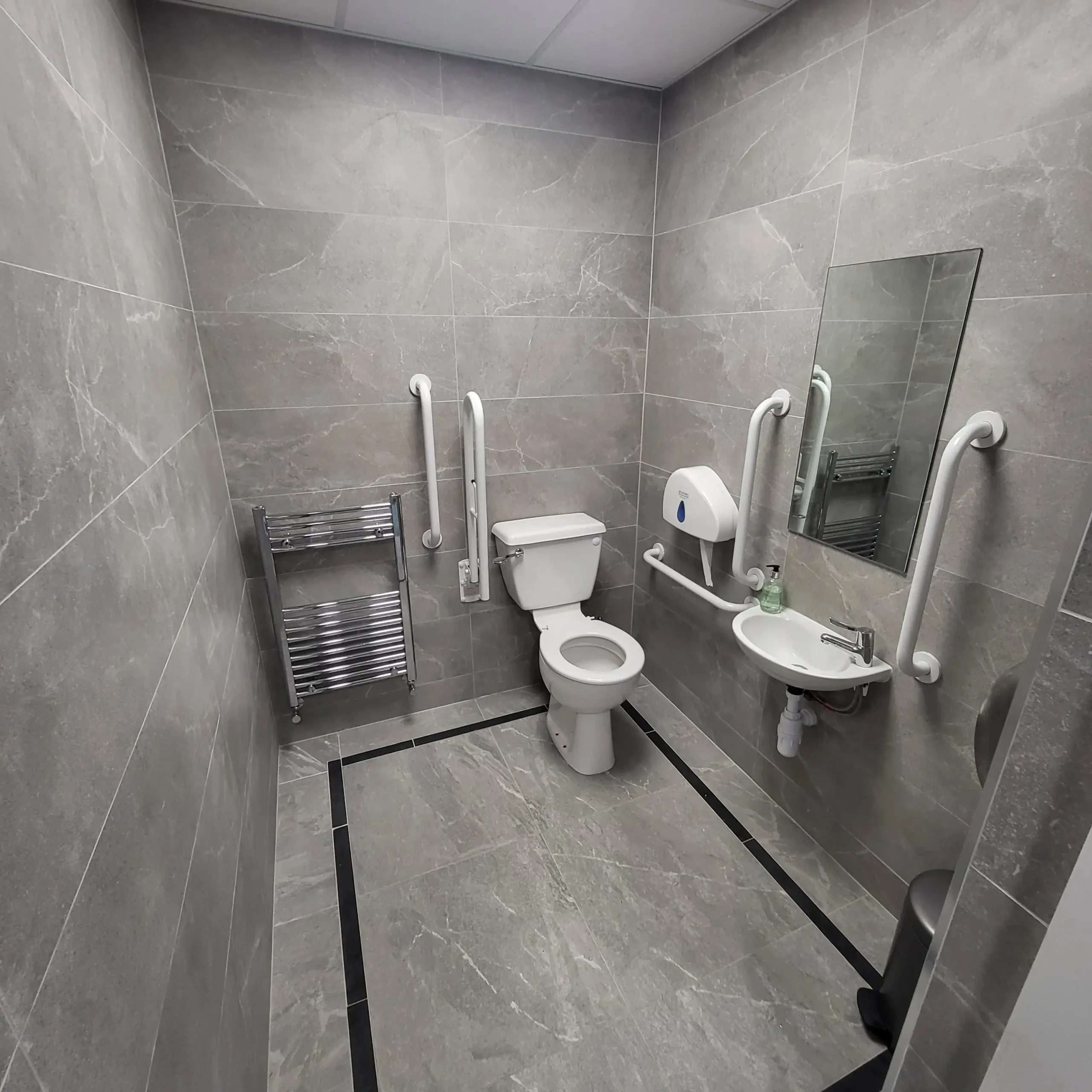 Accessible WC at Sutton Golf Club — grey stone-effect porcelain walls and floor matching the gents zone, wall-hung ceramic basin with white grab rails on either side, close-coupled WC with fixed grab rails, chrome heated towel rail and wall-mounted mirror.